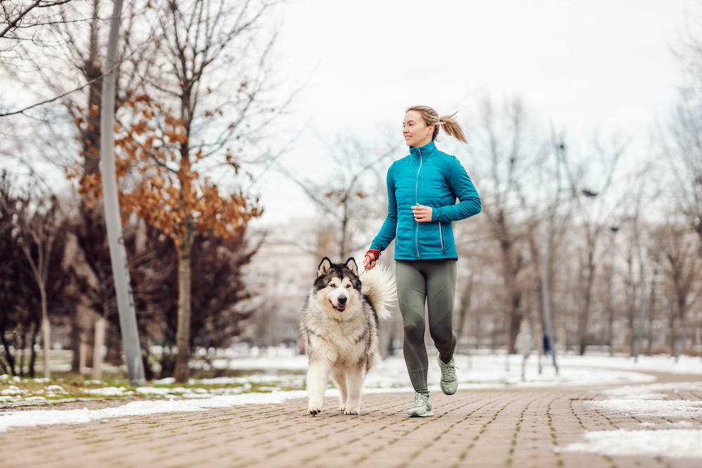 A woman running outdoors through the park with her husky